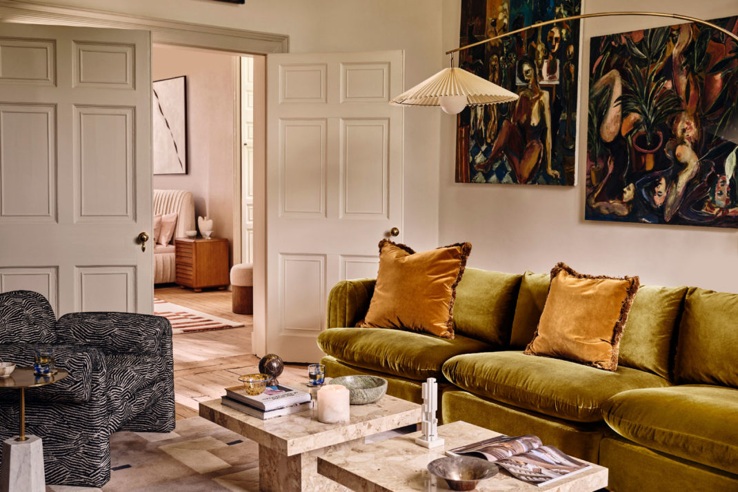 an interior shot of a Soho Home living room, featuring artwork on the walls, a velvety green sofa with orange cushions, and two marbled white coffee tables