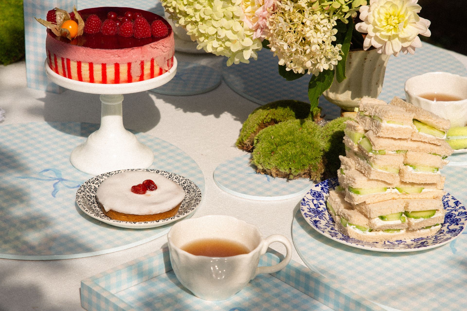 Outdoor table with white tablecloth, blue gingham trays and placemats, and an afternoon tea spread